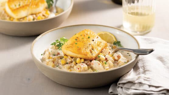 Table set with a white bowl with crab and corn chowder and topped with a pan-seared Chilean Sea Bass. Glass of Chardonnay and another bowl in the background.