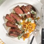 White plate on table with sliced New York strip steak cooked medium-rare and drizzled with garlic aioli and served with hash browns and melted leeks.