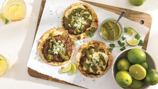 3 Pepper Jack Smash Burger tacos with poblano salsa and sprinkled with cheese on a cutting next to a bowl of limes.