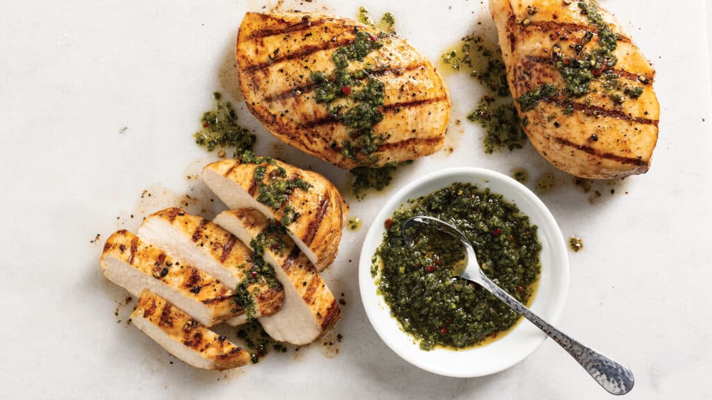 Three grilled chicken breast, one sliced, topped with chimichurri steakhouse topper next to a bowl of chimichurri with a spoon in it.