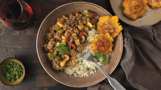 Wood table set with brown bowl with picadillo, white rice and plantains, brown plate with plantains, glass of red wine and small bowl of chopped cilantro