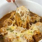Bowl of french onion meatballs with spoon lifting one from the bowl with an epic cheese pull