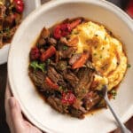 Two hands holding cream-colored bowl with italian pot roast and creamy polenta with au jus. In background is another bowl and a glass of red wine.
