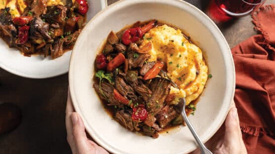 Two hands holding cream-colored bowl with italian pot roast and creamy polenta with au jus. In background is another bowl and a glass of red wine.