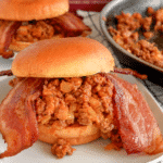 A cheesy bacon and beer sloppy joe with two thick slices of bacon hanging out the bun.