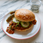 Gochujang sloppy on a sesame seed bun with cucumber slices and pickles on a white plate.