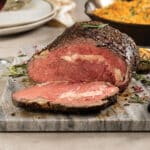 Dijon-rubbed prime rib cooked medium rare and sliced on a marble cutting board. Cast iron skillet with cheddar crumb mashed potatoes, glass of red wine and plates in background.