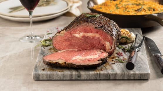 Dijon-rubbed prime rib cooked medium rare and sliced on a marble cutting board. Cast iron skillet with cheddar crumb mashed potatoes, glass of red wine and plates in background.