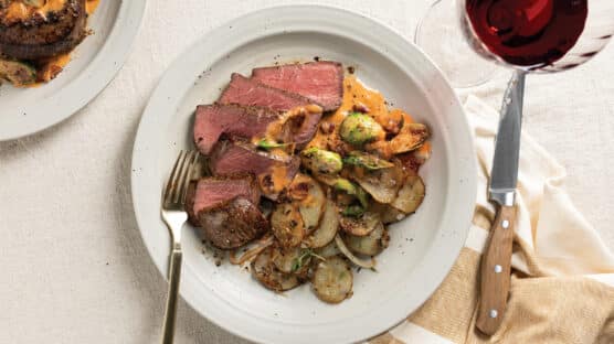 White plate with sliced filet mignon cooked medium rare, Brussels sprouts & bacon with a creamy tomato-white wine sauce, and crispy potatoes and onions.