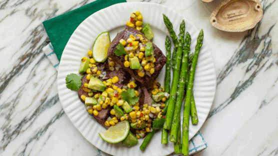 Two grilled Omaha Steaks USDA Certified Tender Private Reserve top sirloin steaks, one whole and one sliced, topped with corn and avocado salsa and served with a side of asparagus, all on a white plate.