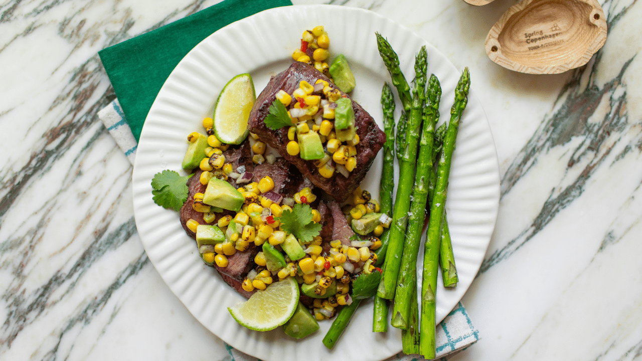 Two grilled Omaha Steaks USDA Certified Tender Private Reserve top sirloin steaks, one whole and one sliced, topped with corn and avocado salsa and served with a side of asparagus, all on a white plate.