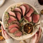 Holiday table with woman holding a cream platter over the table with sliced rosemary-garlic chateaubriand with a small bowl of red wine mushroom sauce. In background on table is a platter of roasted parmesan acorn squash, glass or red wine and white taper candle.