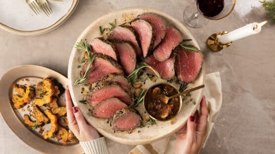 Holiday table with woman holding a cream platter over the table with sliced rosemary-garlic chateaubriand with a small bowl of red wine mushroom sauce. In background on table is a platter of roasted parmesan acorn squash, glass or red wine and white taper candle.