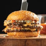 Triple-Decker Buffalo Smash Burger With Blue Cheese Mayo on a wooden cutting board with glass bowl of Buffalo sauce behind it.