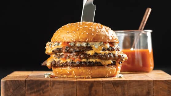 Triple-Decker Buffalo Smash Burger With Blue Cheese Mayo on a wooden cutting board with glass bowl of Buffalo sauce behind it.