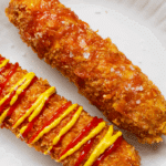 2 Korean-style corn dogs on a white plate. One with ketchup and mustard, one plain.