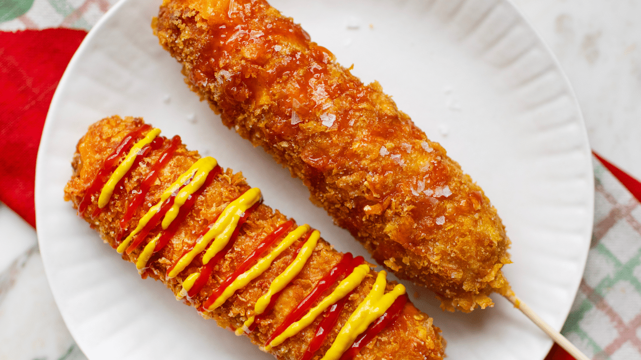 2 Korean-style corn dogs on a white plate. One with ketchup and mustard, one plain.