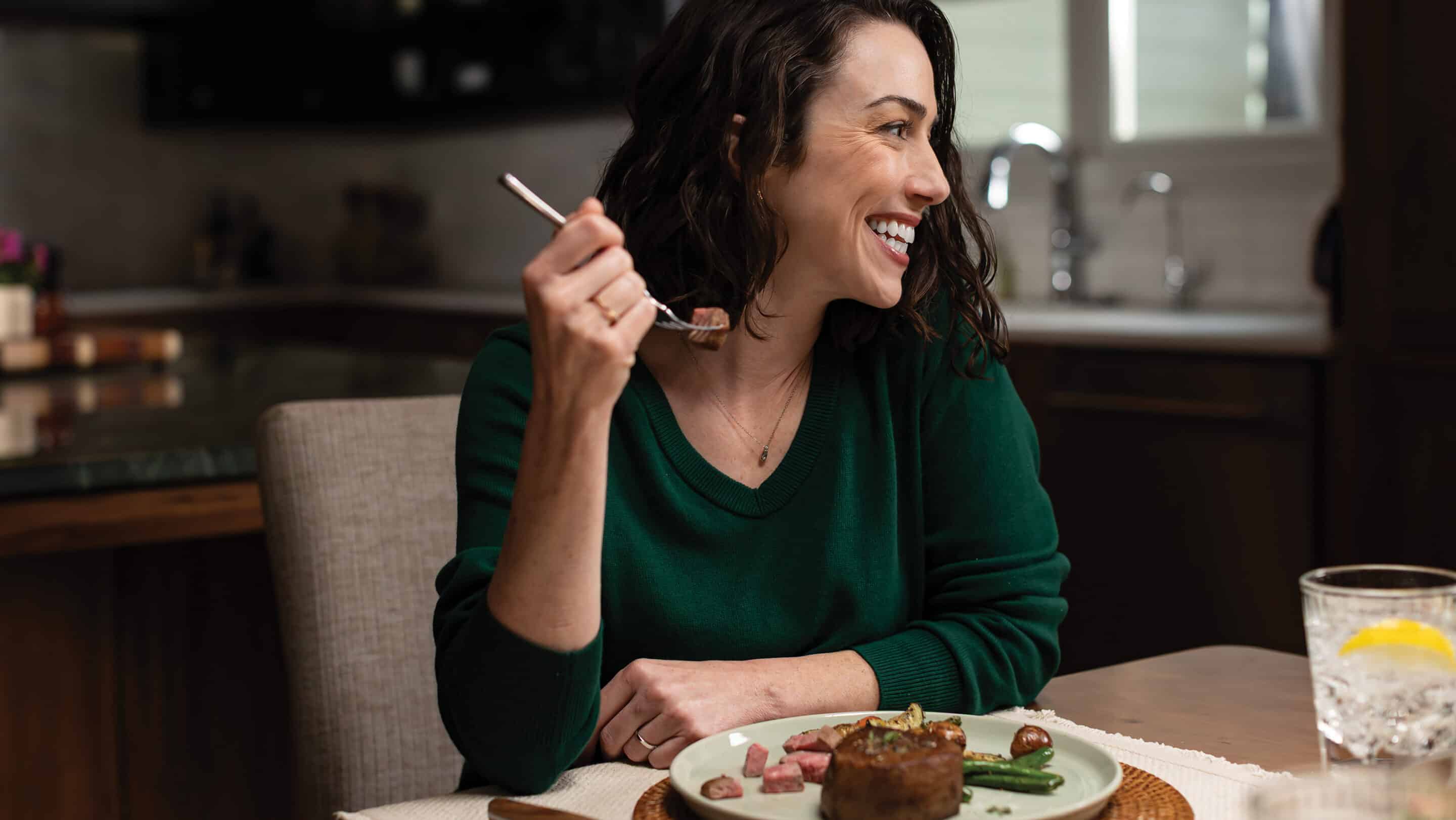 A smiling woman in a green sweater sits at a dining table, holding a fork with a bite of steak while looking off to the side. In front of her is a plate with steak, green beans, and small potatoes, and a glass of water with lemon sits nearby. The setting appears to be a cozy, modern kitchen.