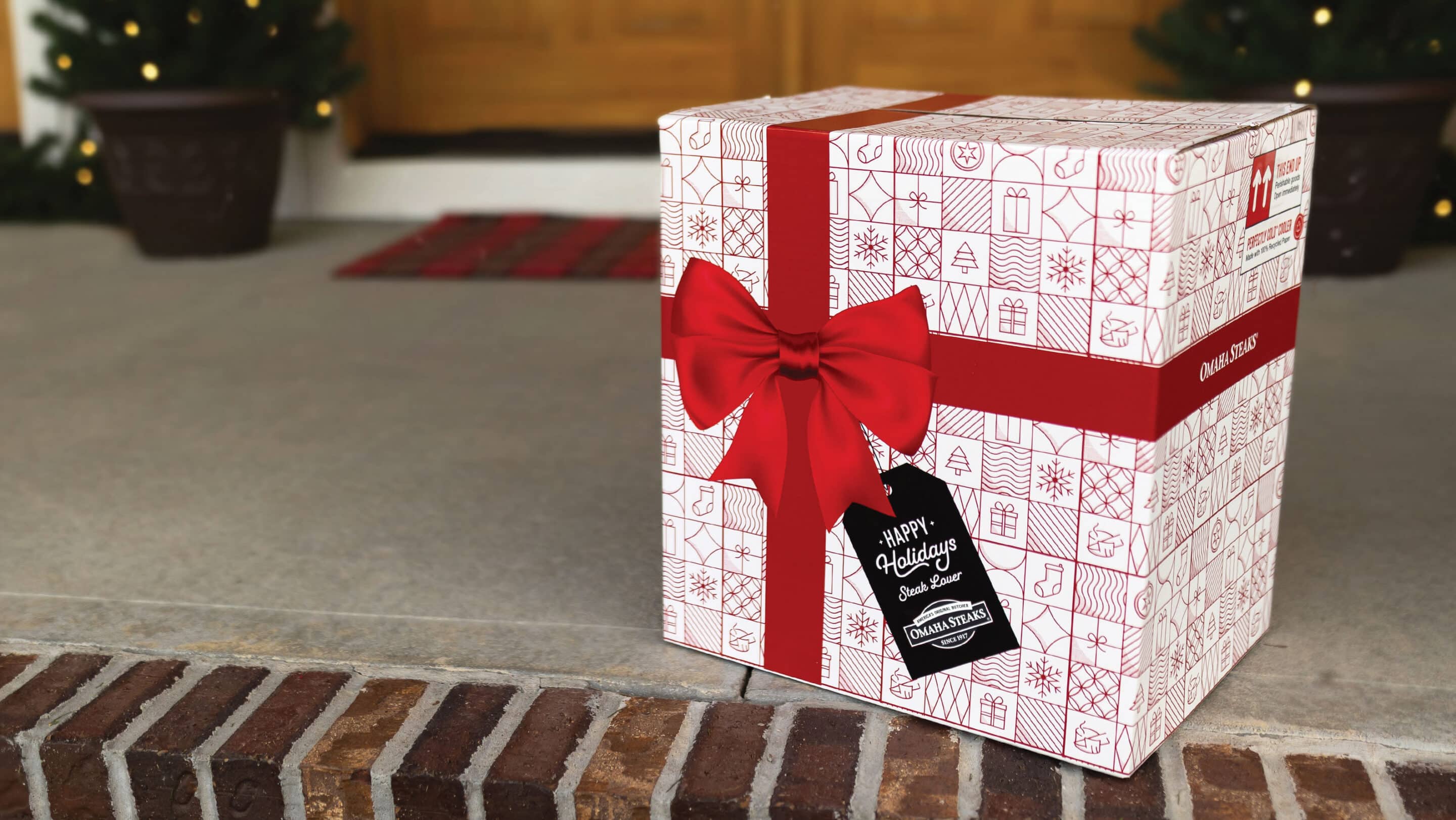 Omaha Steaks holiday gift box with a red bow and “Happy Holidays” gift tag on a front porch.