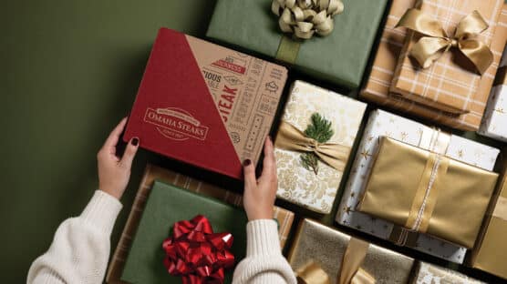 Hands holding an Omaha Steaks gift box surrounded by wrapped holiday presents with colorful bows.