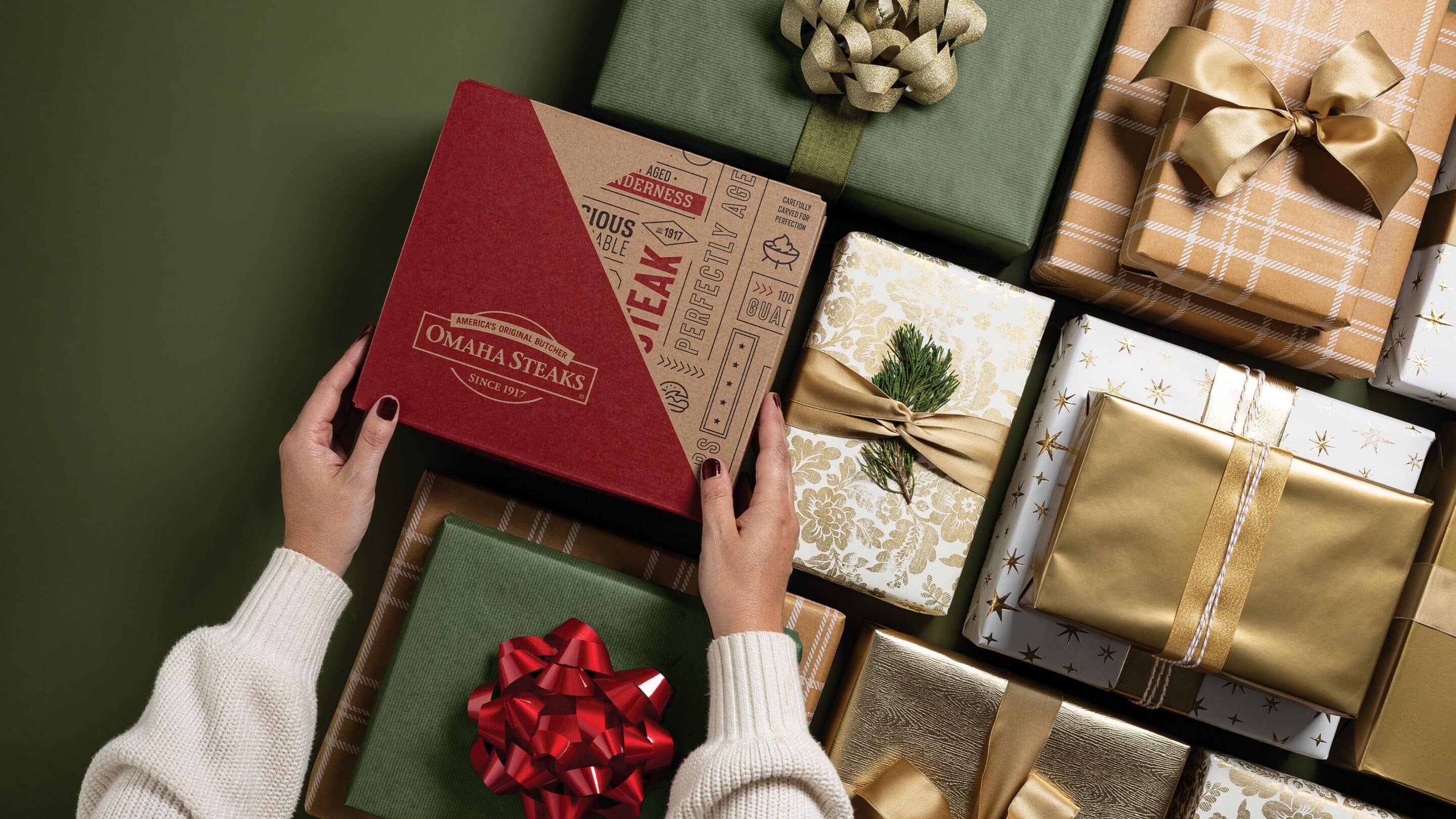 Hands holding an Omaha Steaks gift box surrounded by wrapped holiday presents with colorful bows.
