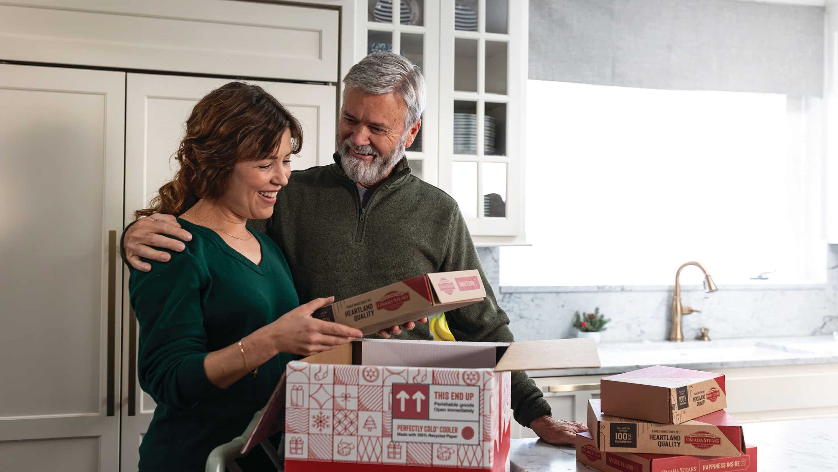 Smiling couple unpacking Omaha Steaks gift boxes together in a bright kitchen.
